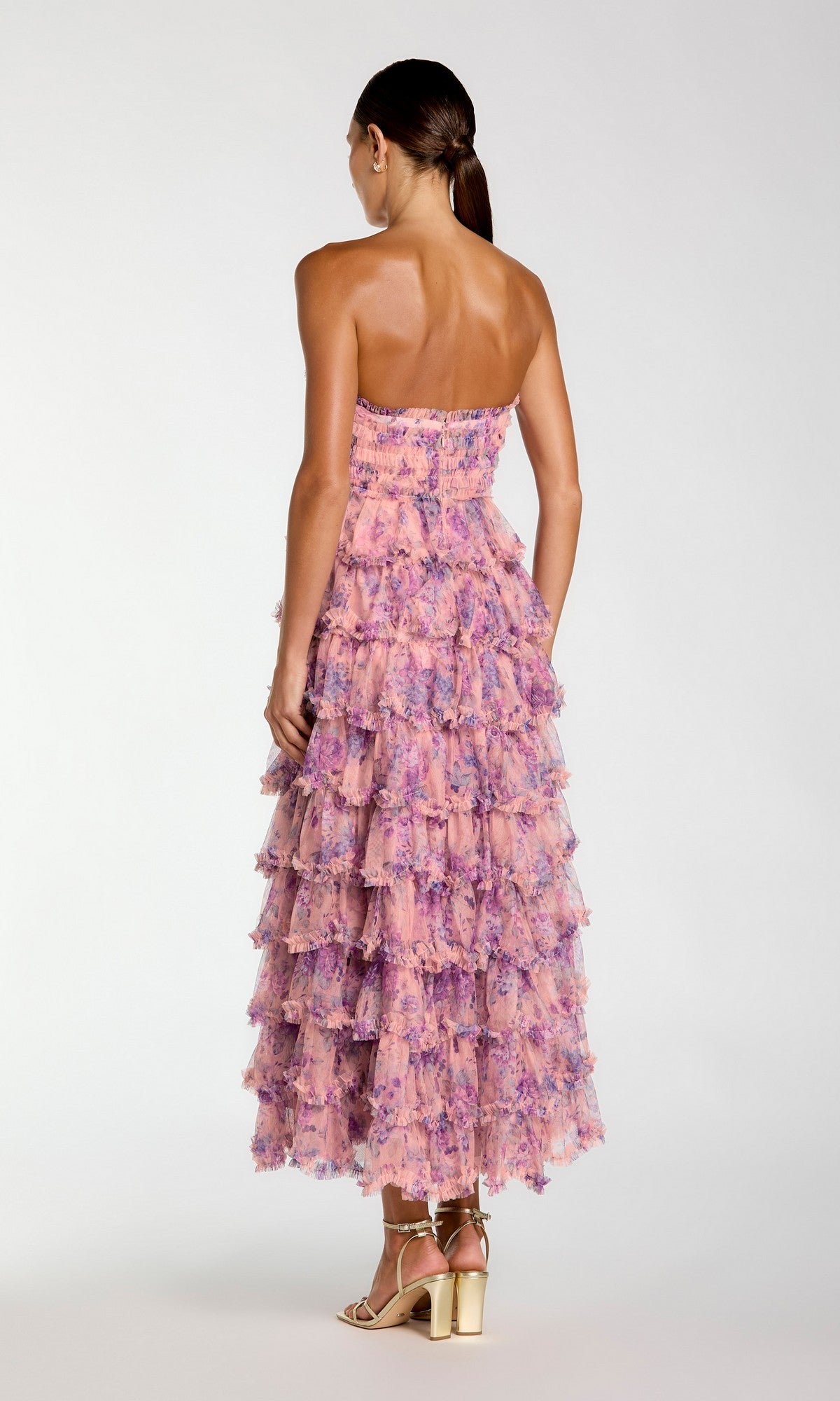 A woman stands with her back to the camera, wearing the Mac Duggal 8420 Tea-Length Party Dress—a strapless midi dress featuring ruffle tiers in pink and purple floral patterns—paired with gold high-heeled sandals against a plain background.